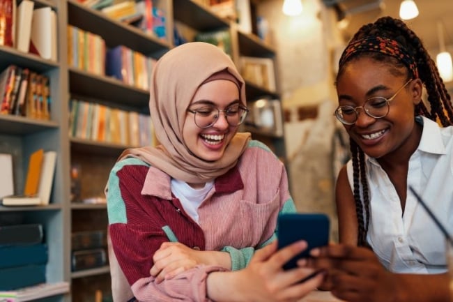 two diverse students sitting in a library are smiling and looking at a phone.